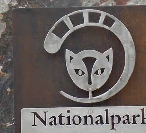 Das Bild zeigt Nationalparklogo. Ein stilisierter Wildkatzenkopf mit dem charakteristischen Schwanz, der von unterhalb des Kopfes in einem Dreiviertelkreis den Kopf umschließt.
