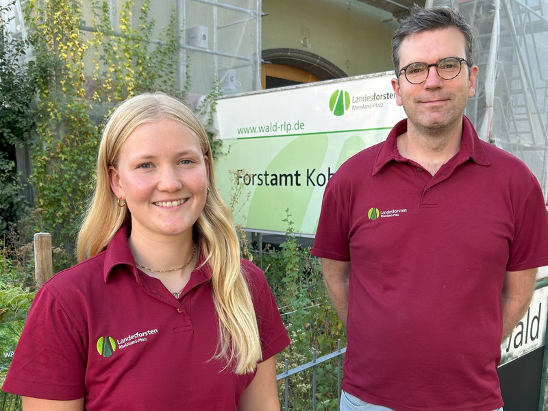 Lina Gilles und Forstamtsleiter Sebastian Schmitz stehen nebeneinander vor dem Forstamtsgebäude. Hinter ihnen ist ein Schild im Erscheinungsbild von Landesforsten Rheinland-Pfalz mit dem Schriftzug Forstamt Koblenz zu erkennen. 