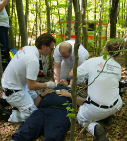 Nach Eintreffen des Rettungsdienstes am Unfallort versorgen drei DRK-Mitarbeiter in weißer Arbeitskleidung kniend das rücklings auf Waldboden liegende Opfer, wobei einer den Kopf des Opfers stabilisierend hält.