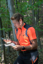 Forstwirt-Azubi Fabian Krämer steht, in orange-schwarzer Arbeitskleidung an einem Baum lehnend, am Unfallort im Wald, das Handy in der rechten, die Rettungskarte in der linken Hand, und setzt, auf diese Hilfsmittel blickend, den Notruf ab.