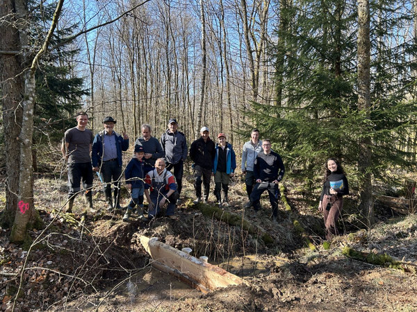 Menschen stehen im Wald und schauen lächelnd in die Kamera. Im Vordergrund ein historischer Entwässerungsgraben mit frisch eingebauter Spundwand aus heimischem Holz.