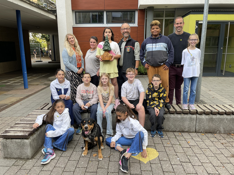 In drei Reihen haben sich die Kinder der Klasse 5b "aufgestellt". Ganz vorne sitzen zwei Schülerinnen auf dem Boden, dazwischen der Hund Balu. In der zweiten Reihe sitzen fünf Kinder auf einer Bank. Dahinter stehen die drei restlichen Kinder flankiert von Förster Ebi und drei Erwachsenen vom Lehrerkollegium.